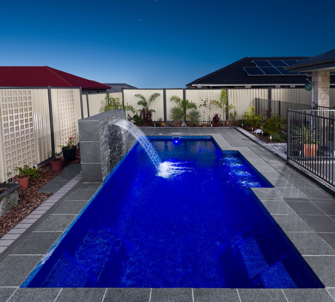A rectangular fibreglass pool with a cascading water blade and blue-lit water at dusk, surrounded by granite-topped paving and landscaped garden in a suburban Brisbane, QLD backyard