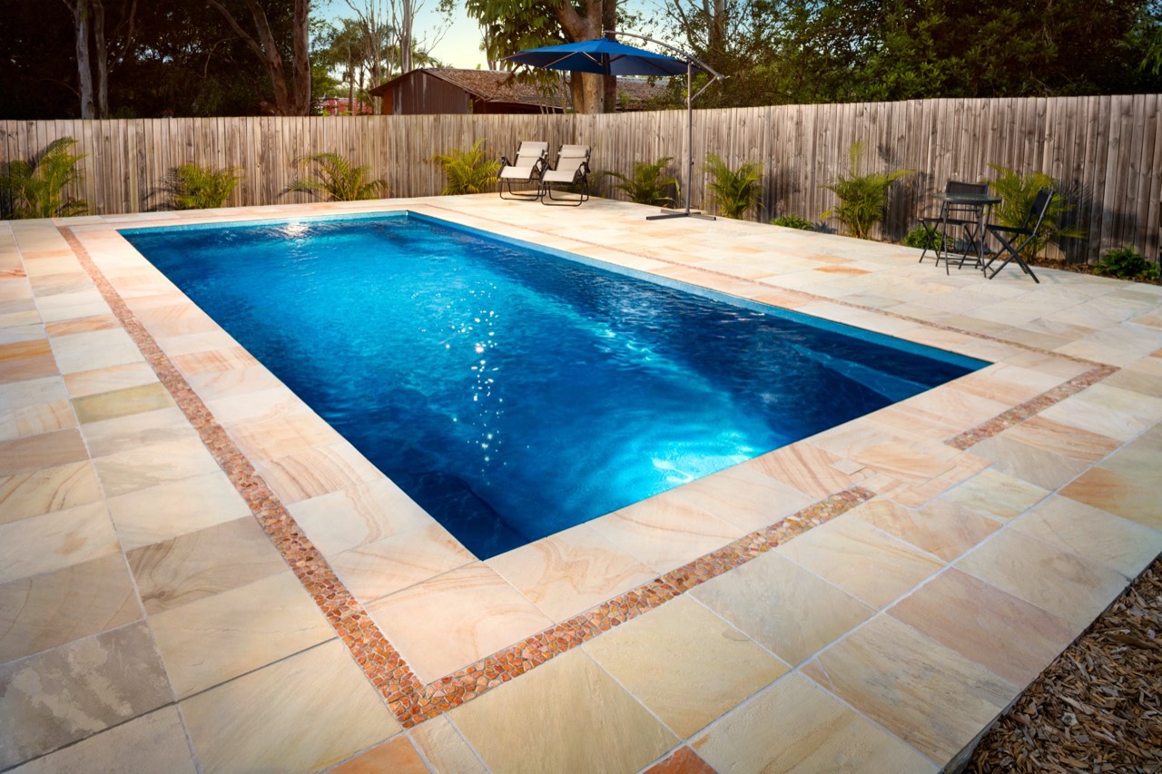 A rectangular fibreglass pool with sandstone paving and a decorative tiled border in a fenced Brisbane, QLD backyard