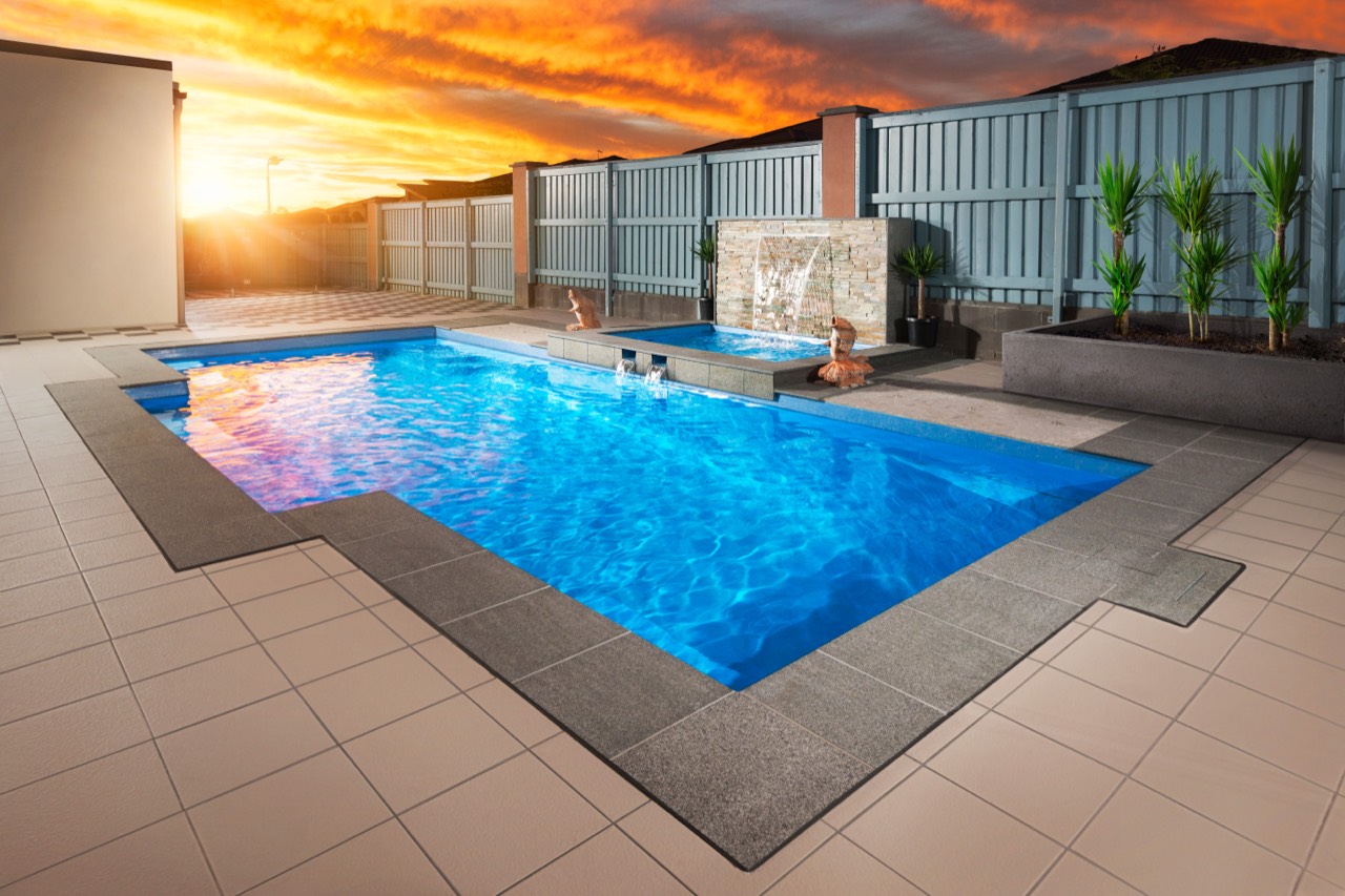 A fibreglass pool with a feature water blade at dusk, lit by a vivid sunset over a fenced Brisbane, QLD backyard